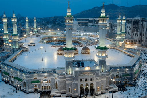 An aerial view of the Masjid al-Haram in Mecca, Saudi Arabia, at dusk, showing the Kaaba in the center surrounded by a vast number of pilgrims performing the Tawaf